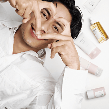 Hudson Williams smiles and forms a heart shape with his hands in front of his face, with skincare products scattered around him.